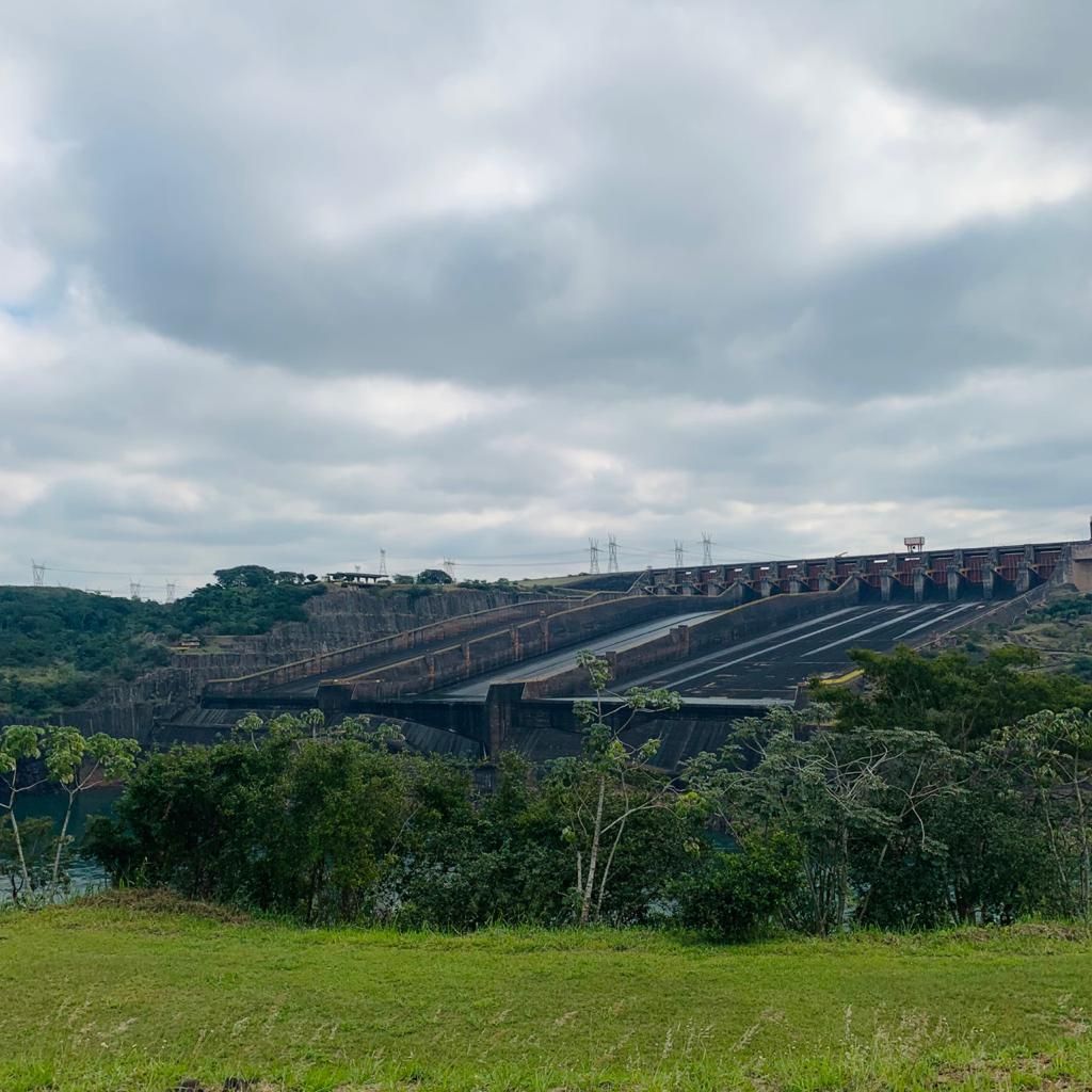 Grande barragem de concreto com vertedouros sob um céu nublado. Vegetação verdejante em primeiro plano.