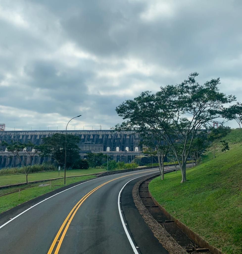 Uma estrada sinuosa leva a uma grande represa sob um céu nublado. Grama verde e árvores ladeiam a estrada.