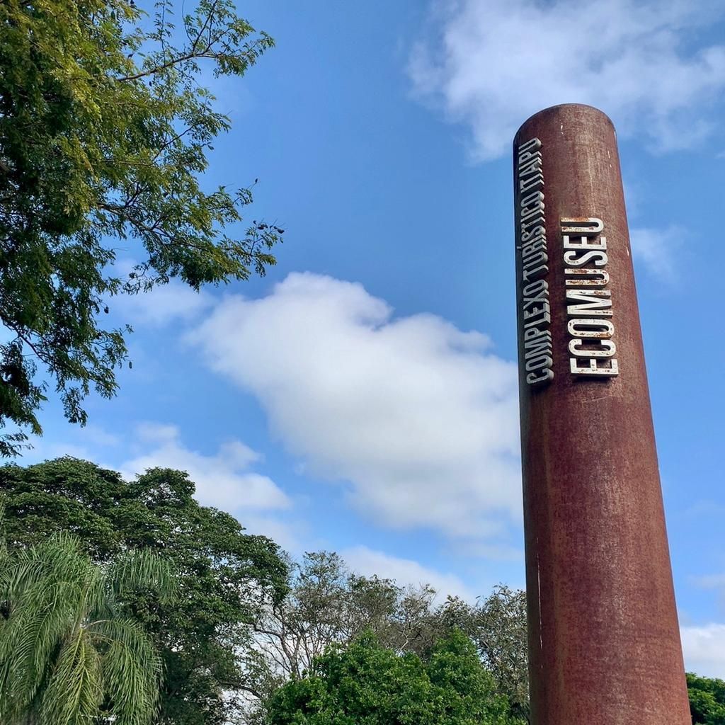Placa alta marrom enferrujada do Ecomuseu contra um céu azul com nuvens e árvores.