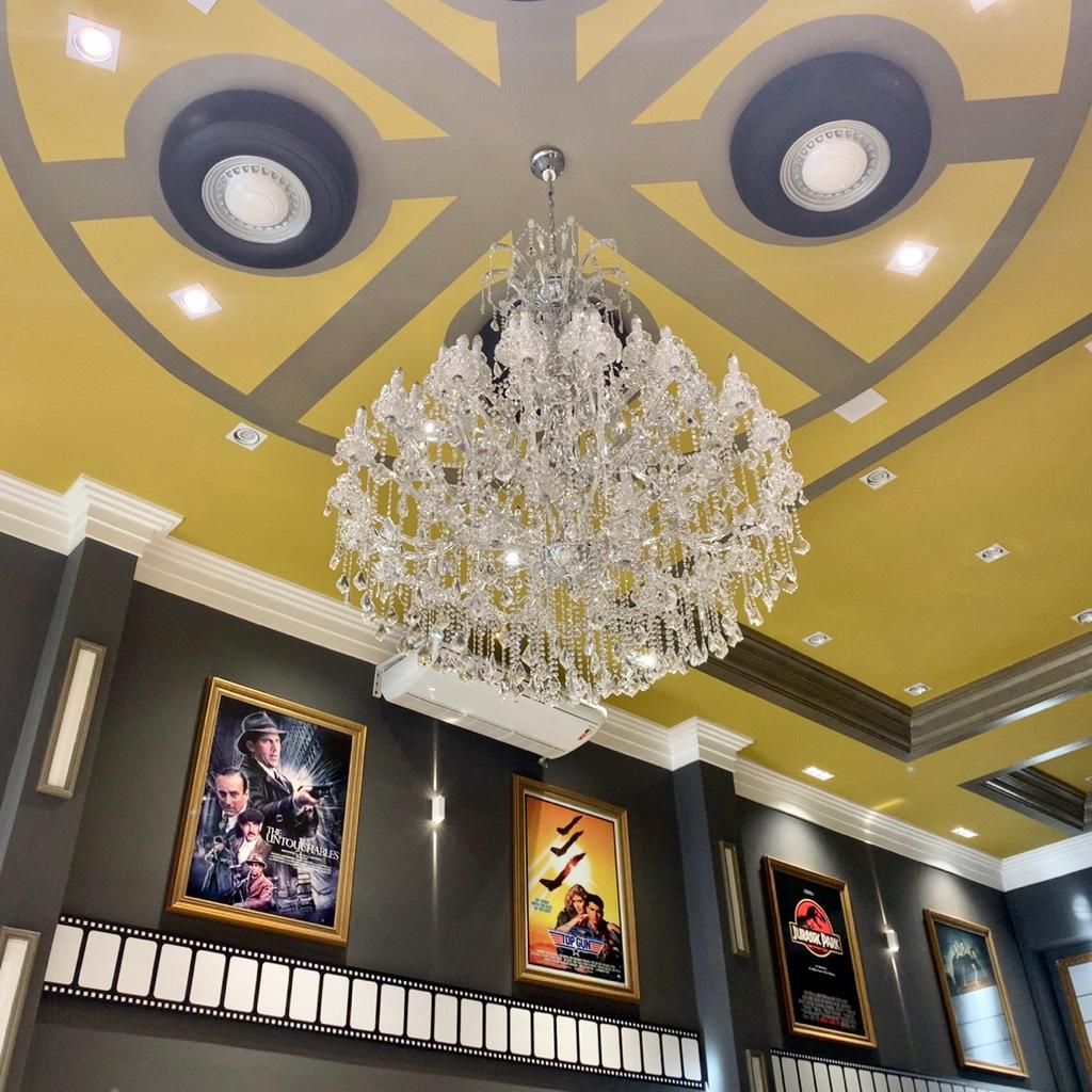Lustre de cristal pendurado em um cinema, teto amarelo, pôsteres de filmes em parede cinza escuro.