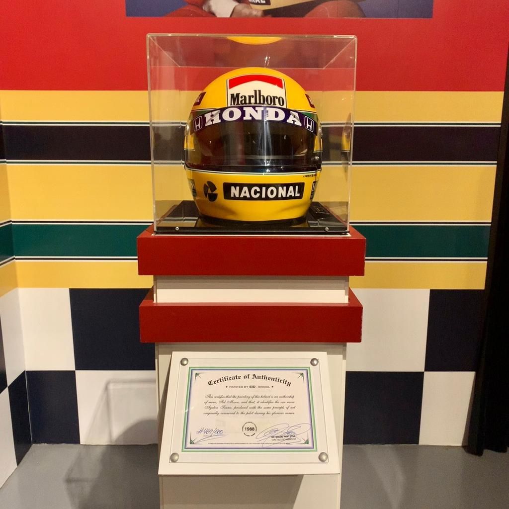 Capacete amarelo, vermelho e branco em vitrine com certificado. Marcas Marlboro, Honda e Nacional.