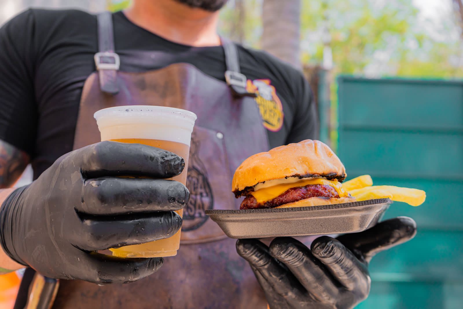 Pessoa usando luvas pretas segurando hambúrguer e cerveja ao ar livre.