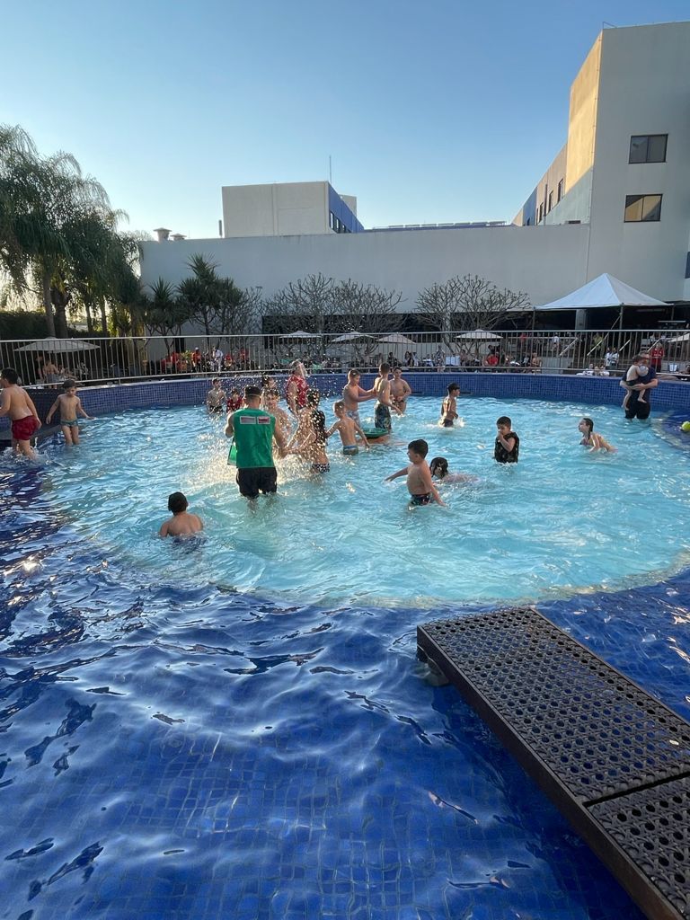 Pessoas brincando em uma piscina de um resort, água azul, dia ensolarado, prédio branco ao fundo.