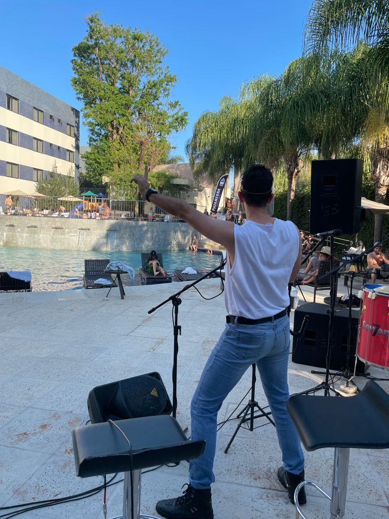 Homem no palco se apresentando com microfone, piscina e prédio ao fundo.