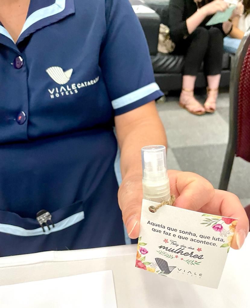 Mulher de uniforme azul segurando um pequeno frasco de spray com uma etiqueta. A etiqueta tem texto e detalhes florais.