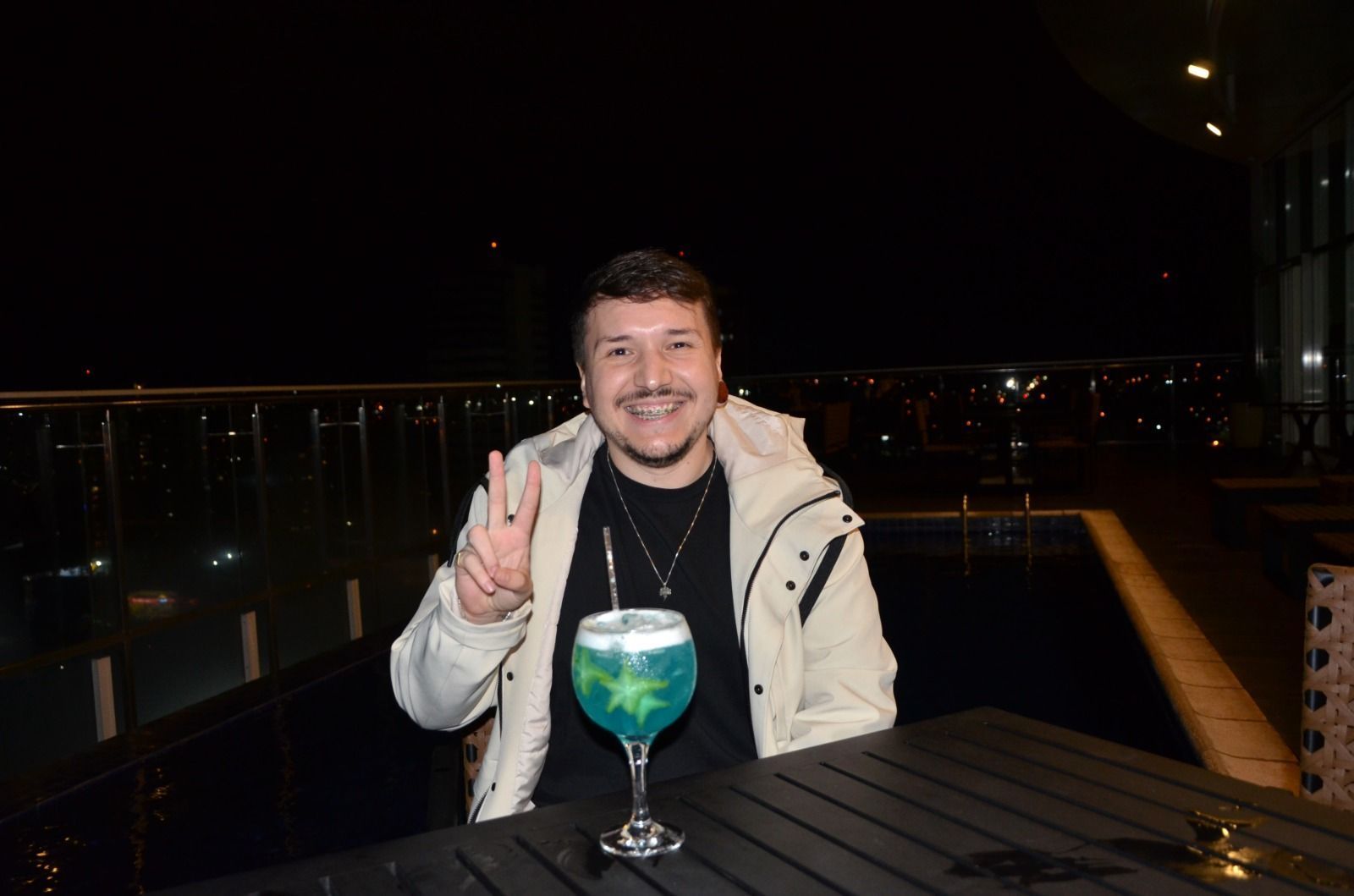 Homem à noite, sorrindo, fazendo o sinal da paz. Sentado com um coquetel azul no terraço com vista para a cidade.
