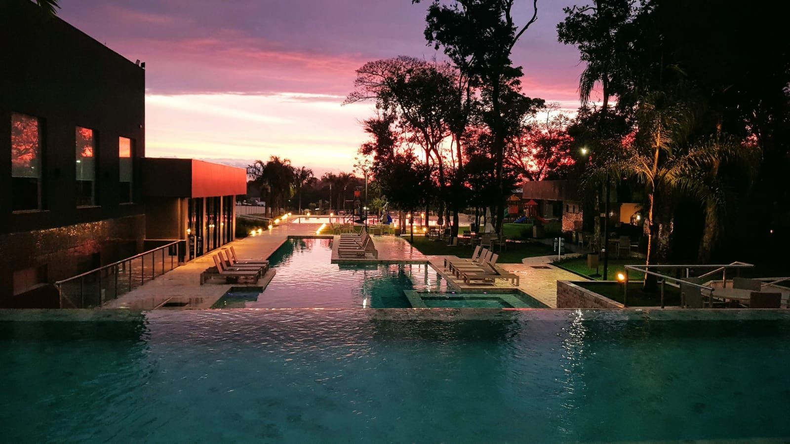 Pôr do sol sobre a piscina de um resort, com céu rosa e laranja, prédios, árvores e espreguiçadeiras.