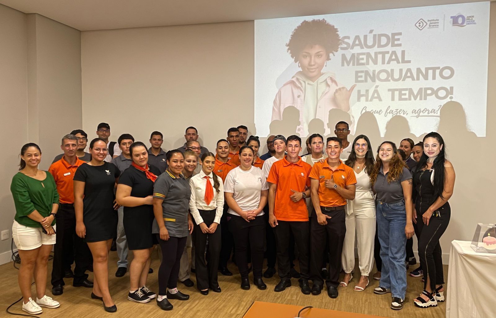 Grupo de pessoas posando em frente a uma tela com texto sobre saúde mental. Em ambiente interno.