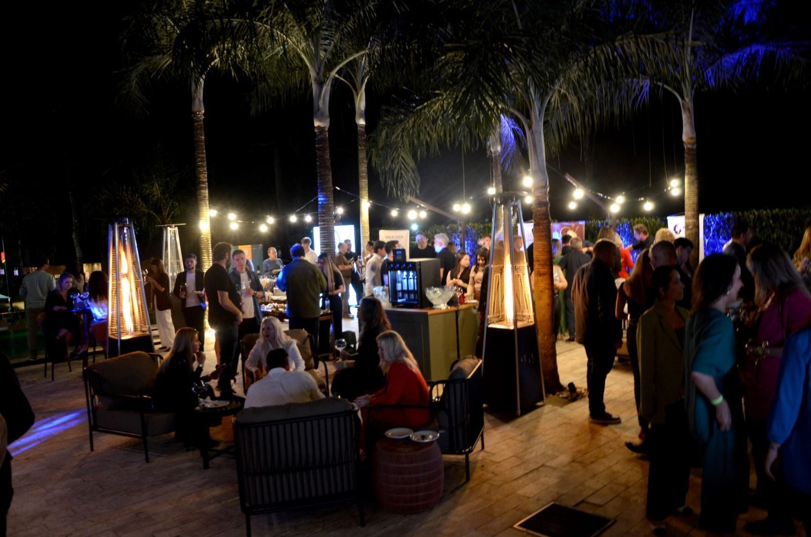 Evento noturno ao ar livre: pessoas socializando sob luzes de corda e palmeiras. Aquecedores de lareira, bebidas e comidas estão disponíveis.