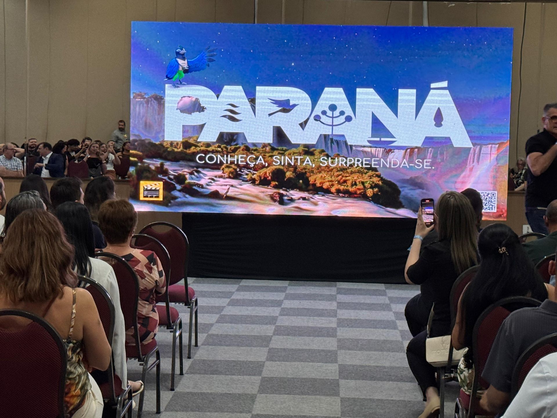 O público assiste a uma apresentação com uma tela exibindo