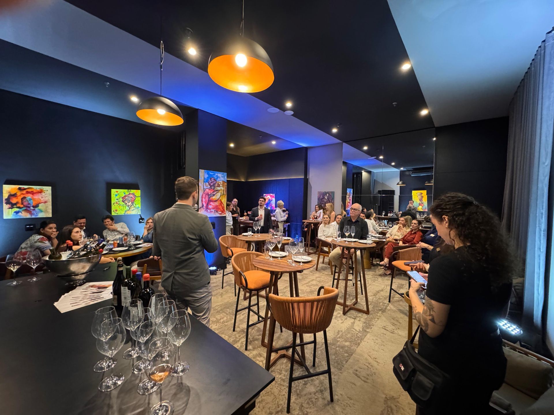 Pessoas reunidas em uma sala moderna, ouvindo um palestrante, taças de vinho em um bar, pinturas nas paredes, iluminação azul e laranja.