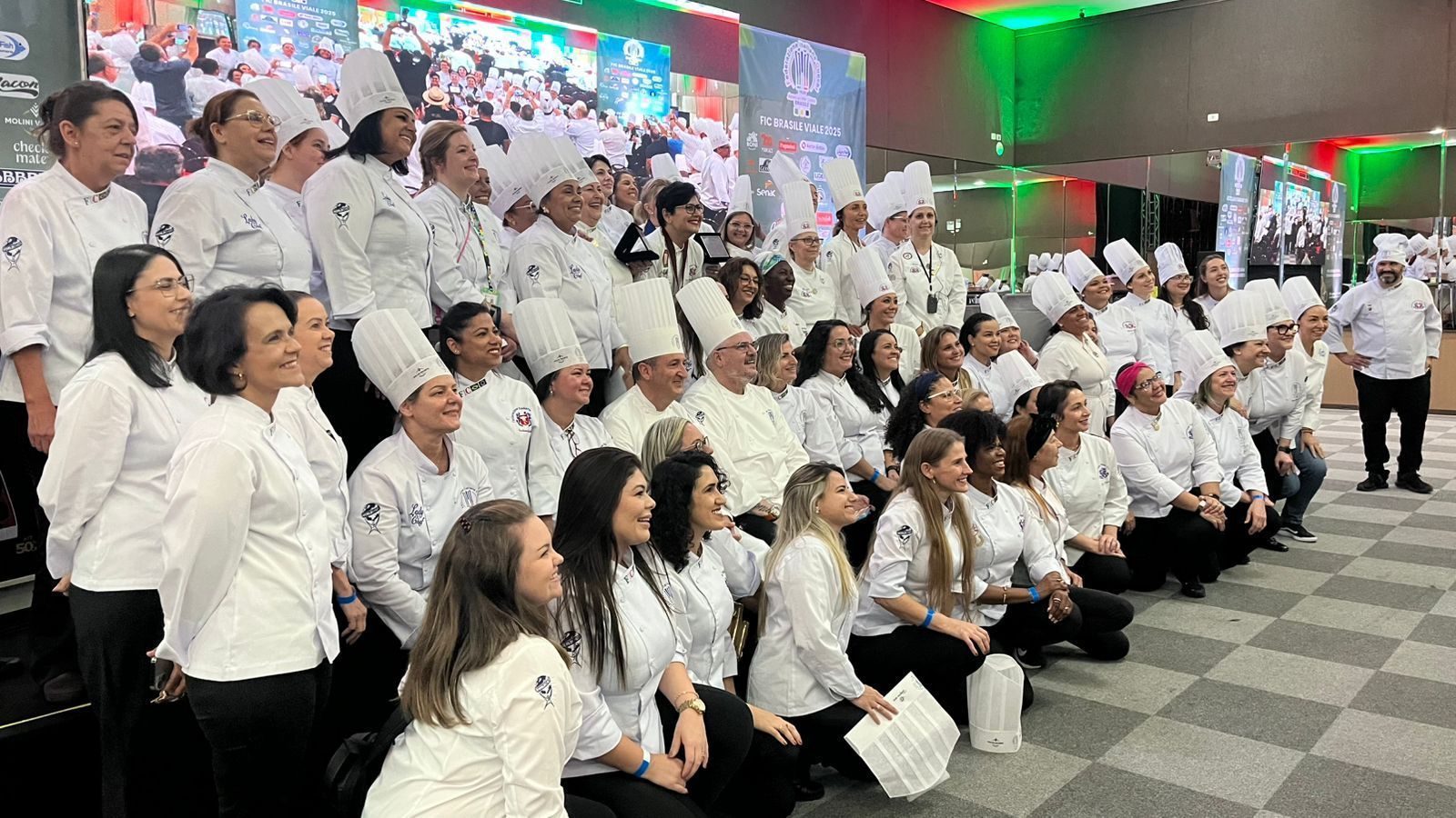 Grupo de chefs em uniformes brancos, posando juntos em um ambiente interno sob iluminação festiva.