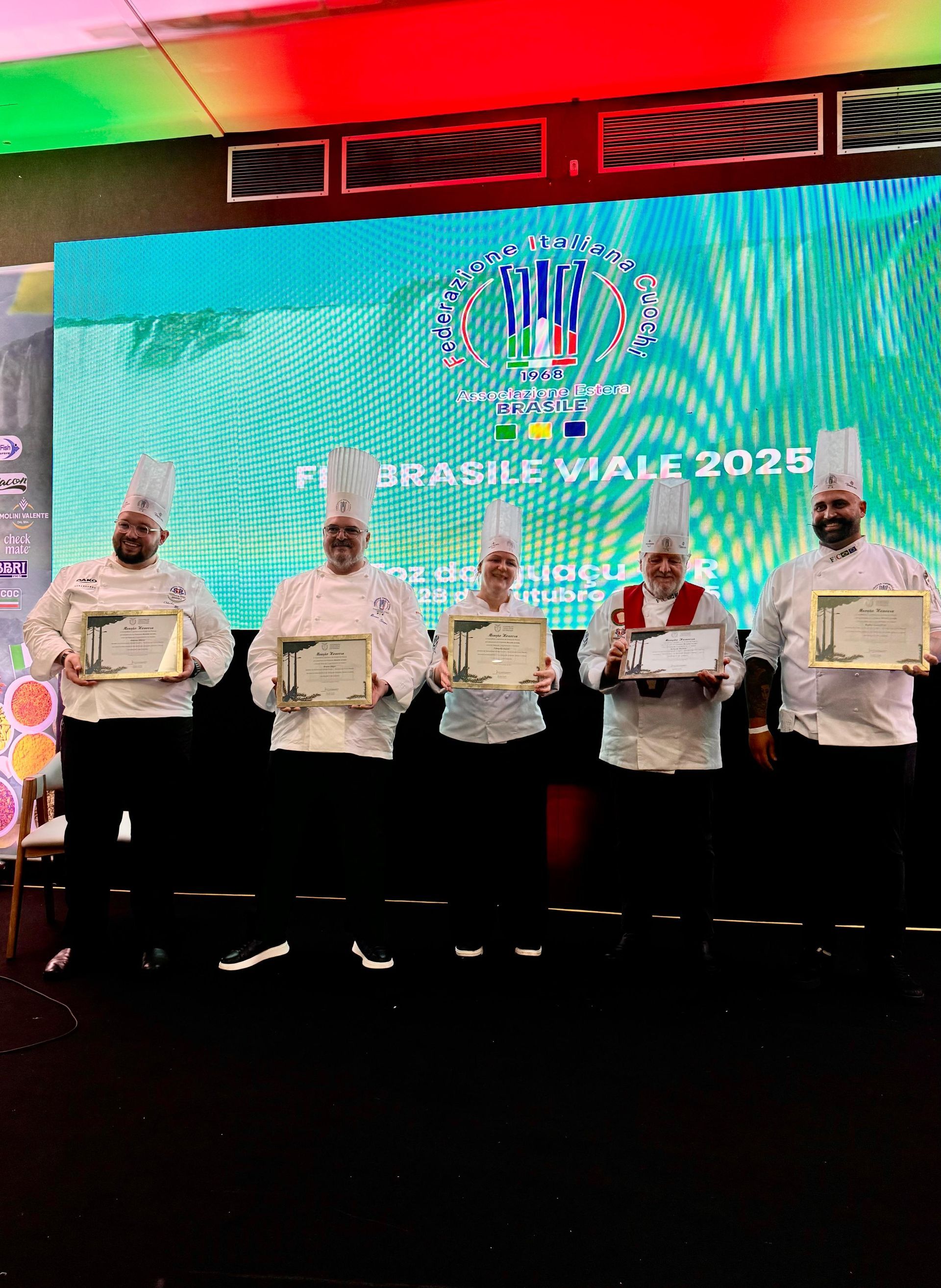 Cinco chefs de uniforme branco seguram certificados emoldurados em um palco com uma tela exibindo um logotipo.