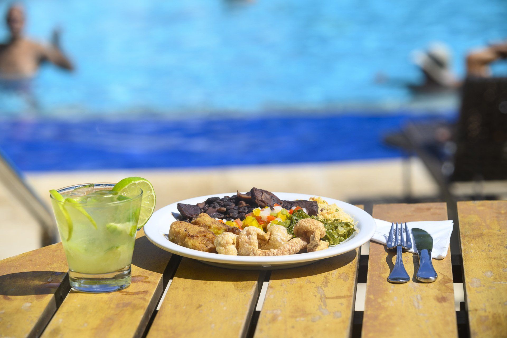 Prato de comida e bebida em uma mesa perto da piscina, com pessoas nadando ao fundo.