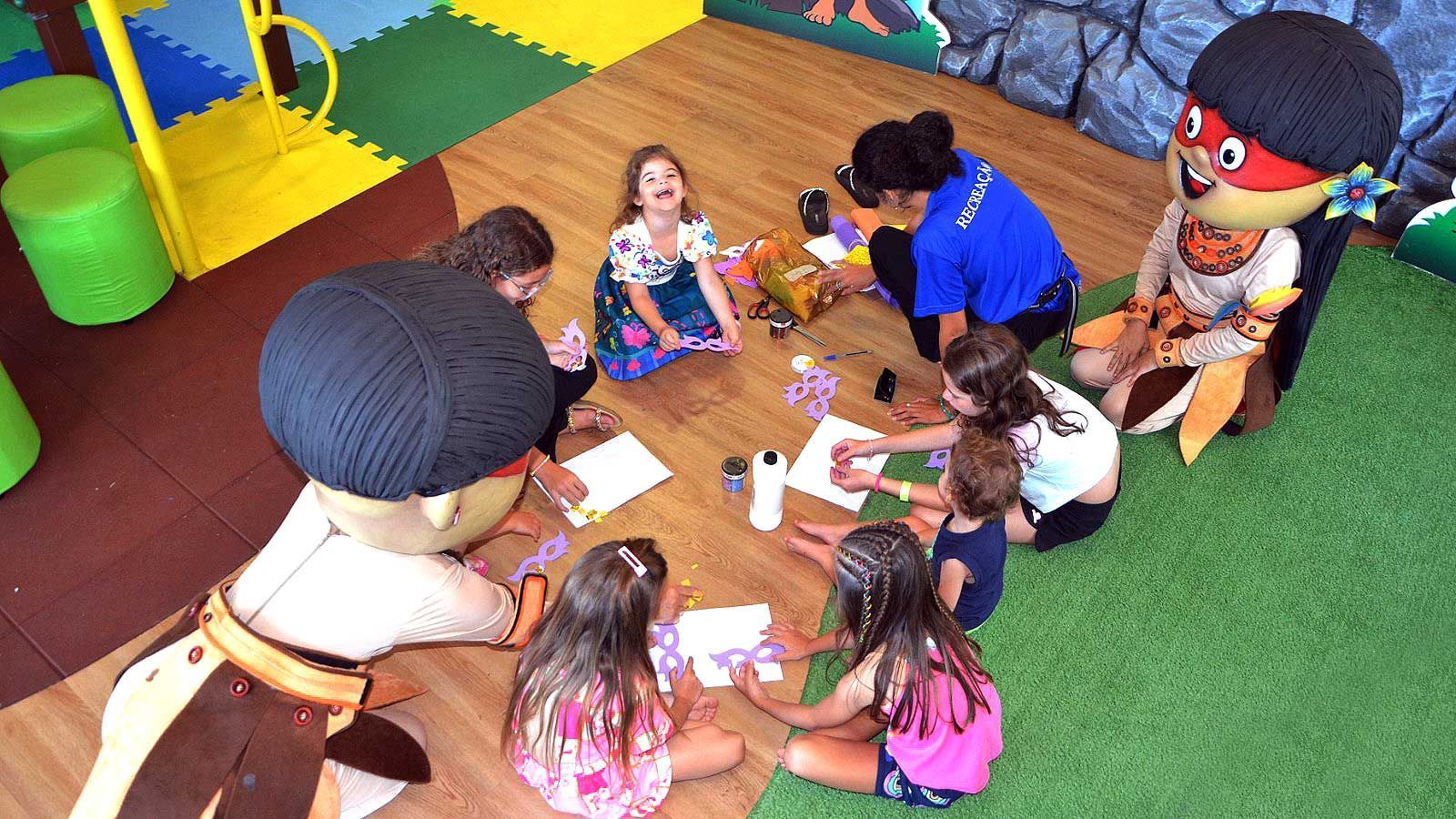 Crianças e adultos em uma sala de jogos, colorindo em uma mesa. Um mascote está por perto.