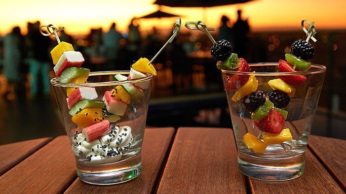 Dois copos com espetos de frutas sobre uma mesa de madeira, com vista para uma festa no terraço ao pôr do sol.