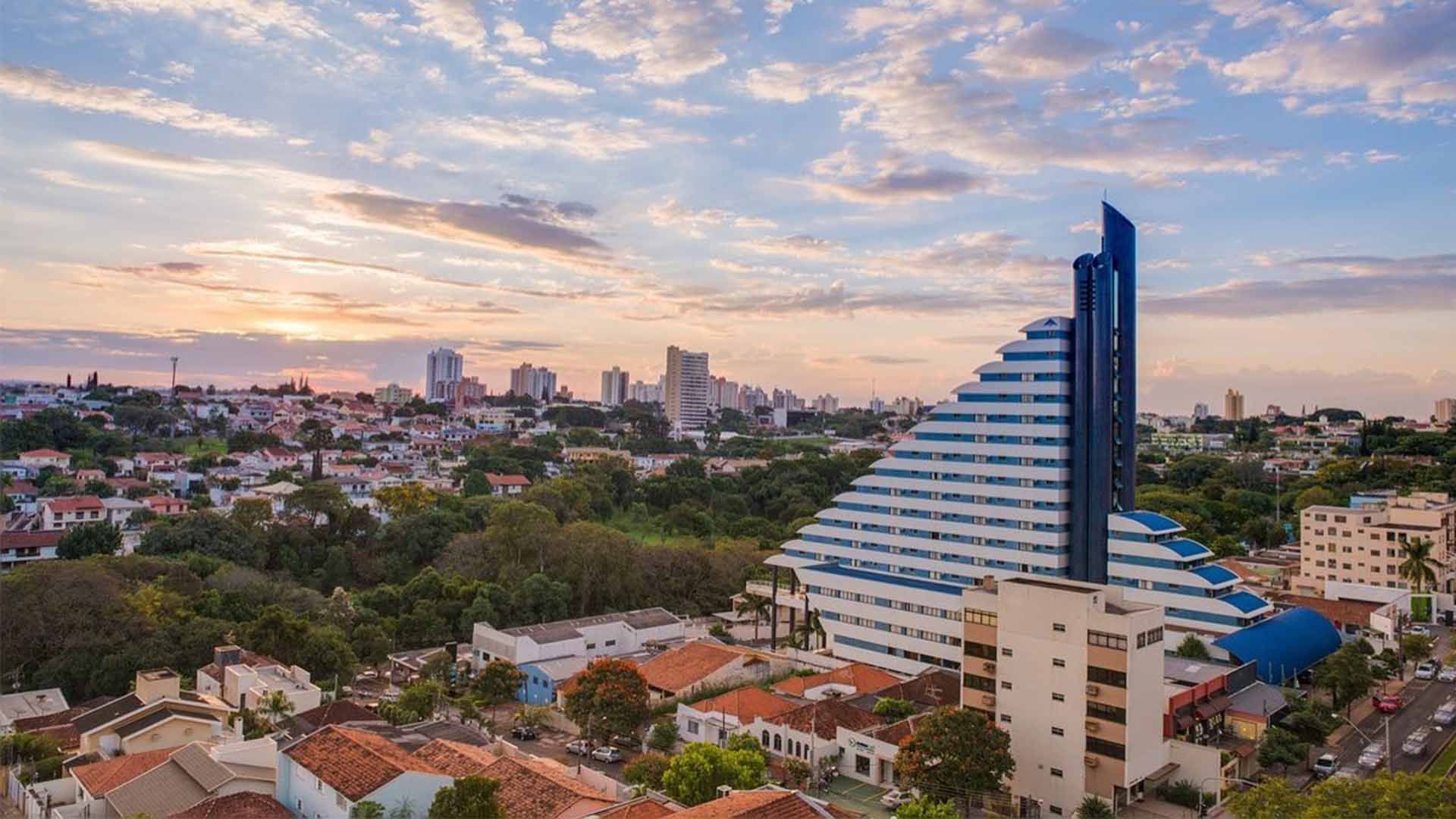 Vista da cidade ao pôr do sol, com um edifício azul e branco de vários andares em destaque no primeiro plano.