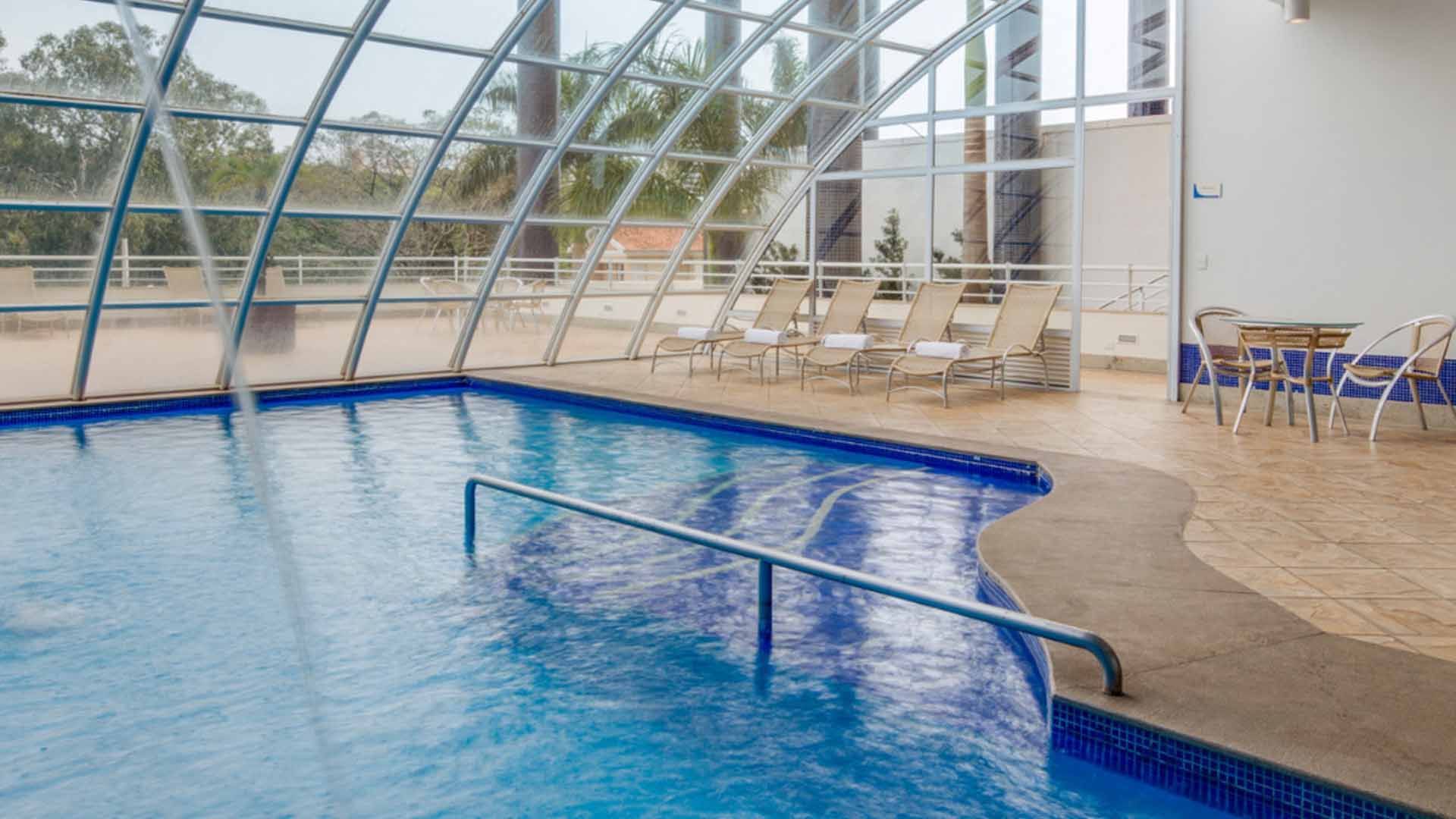 Grande edifício azul e branco, provavelmente um hospital ou escritório, cercado por estradas, árvores e carros estacionados.