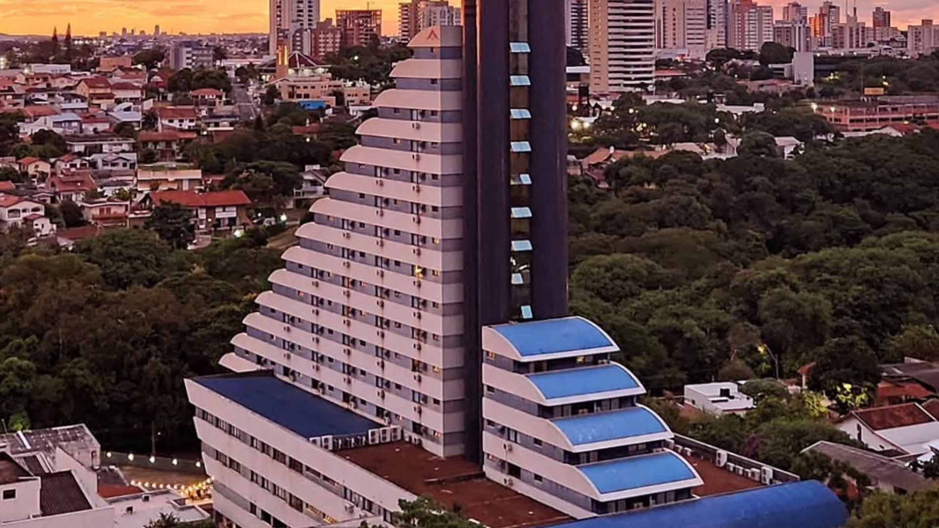 Um prédio alto, branco e escalonado, com uma torre central escura e detalhes em azul, situado em uma cidade com vegetação e outros edifícios.