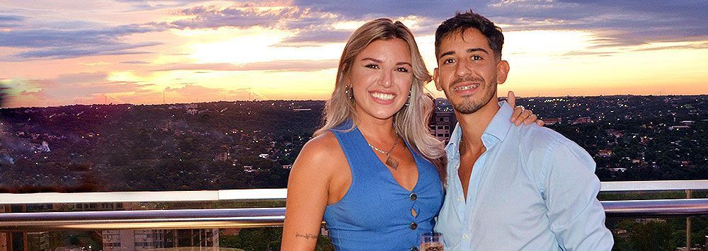 Um casal sorridente posa em uma sacada enquanto o sol se põe sobre uma paisagem urbana.