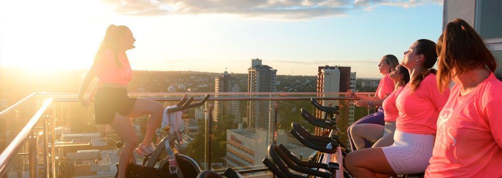 Pessoas com camisas rosas pedalam no telhado de uma casa, com o horizonte da cidade ao fundo, ao pôr do sol.