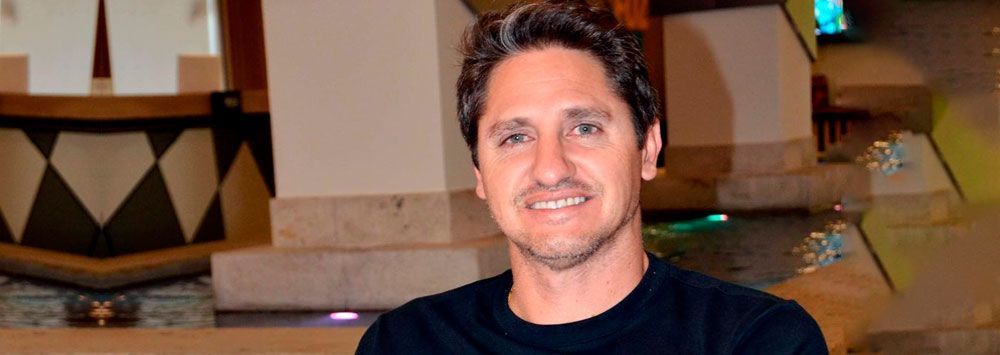 Um homem sorridente, com cabelos grisalhos e uma camisa escura, está em um ambiente interno com um muro de pedra e uma coluna decorativa ao fundo.