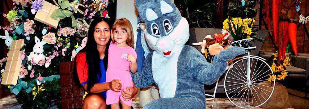 Uma mulher e uma menina posam com o Pernalonga; há uma árvore de flores com um monociclo ao fundo.