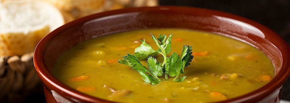Uma tigela de sopa de ervilha, decorada com coentro e pão crocante ao fundo.