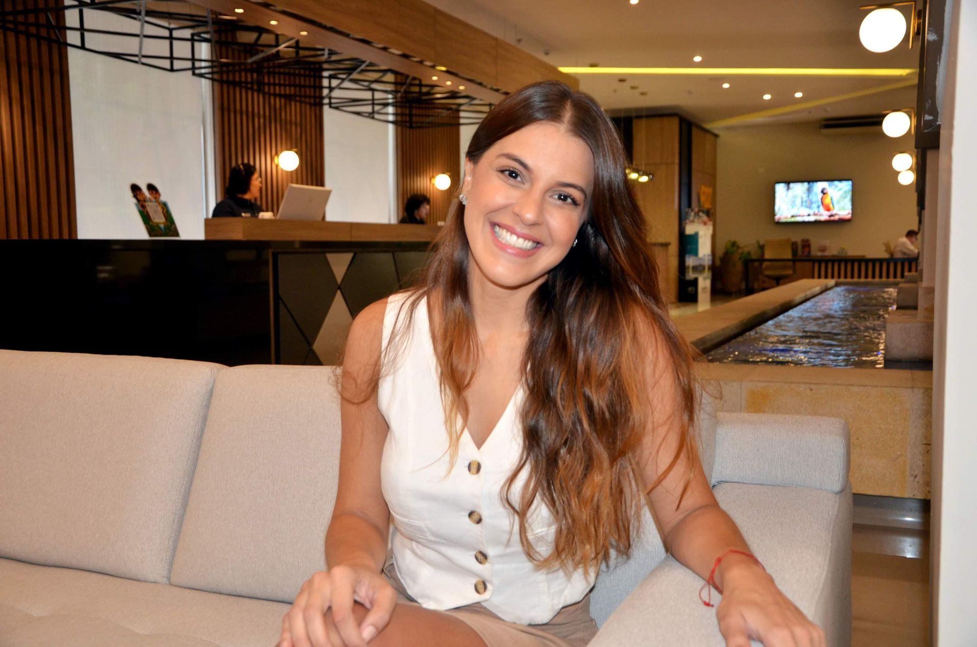Mulher de cabelos castanhos sorrindo, sentada em um sofá bege no saguão de um hotel moderno.
