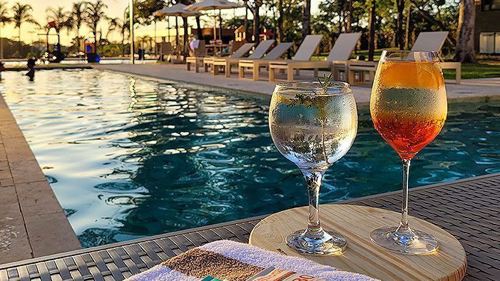 Dois coquetéis em uma bandeja de madeira perto de uma piscina, espreguiçadeiras e guarda-sóis.
