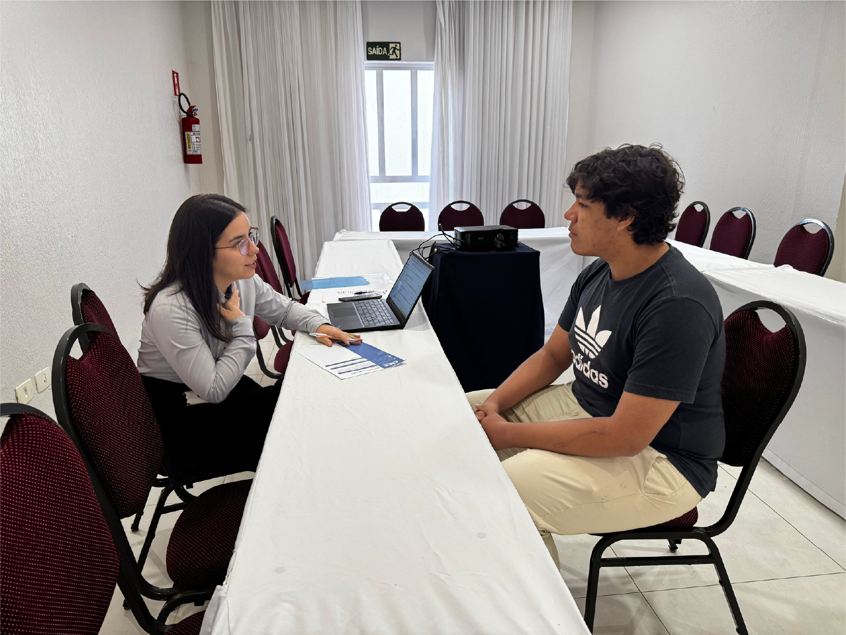 Mulher e homem sentados à mesa, conversando. Laptop e papéis presentes. Sala branca, cadeiras vermelhas.