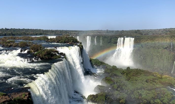 Cataratas do Iguaçu com várias cachoeiras caindo sobre penhascos, um arco-íris visível e vegetação exuberante.