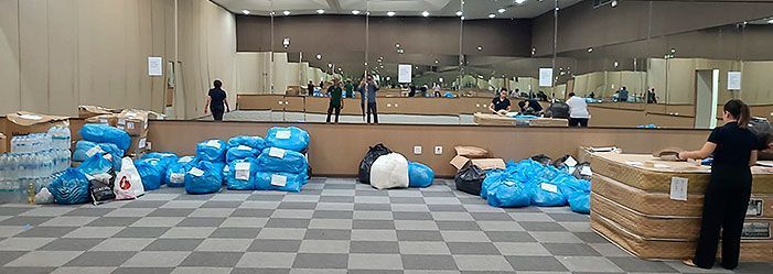 Armazém com pessoas separando doações, pilhas de sacolas azuis e caixas em um chão xadrez.
