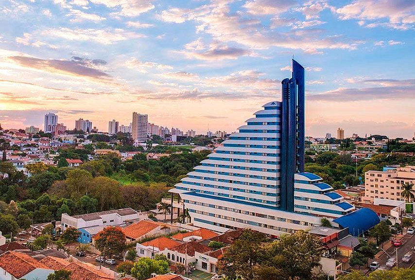 Um prédio alto, branco e escalonado, com uma torre central escura e detalhes em azul, situado em uma cidade com vegetação e outros edifícios.