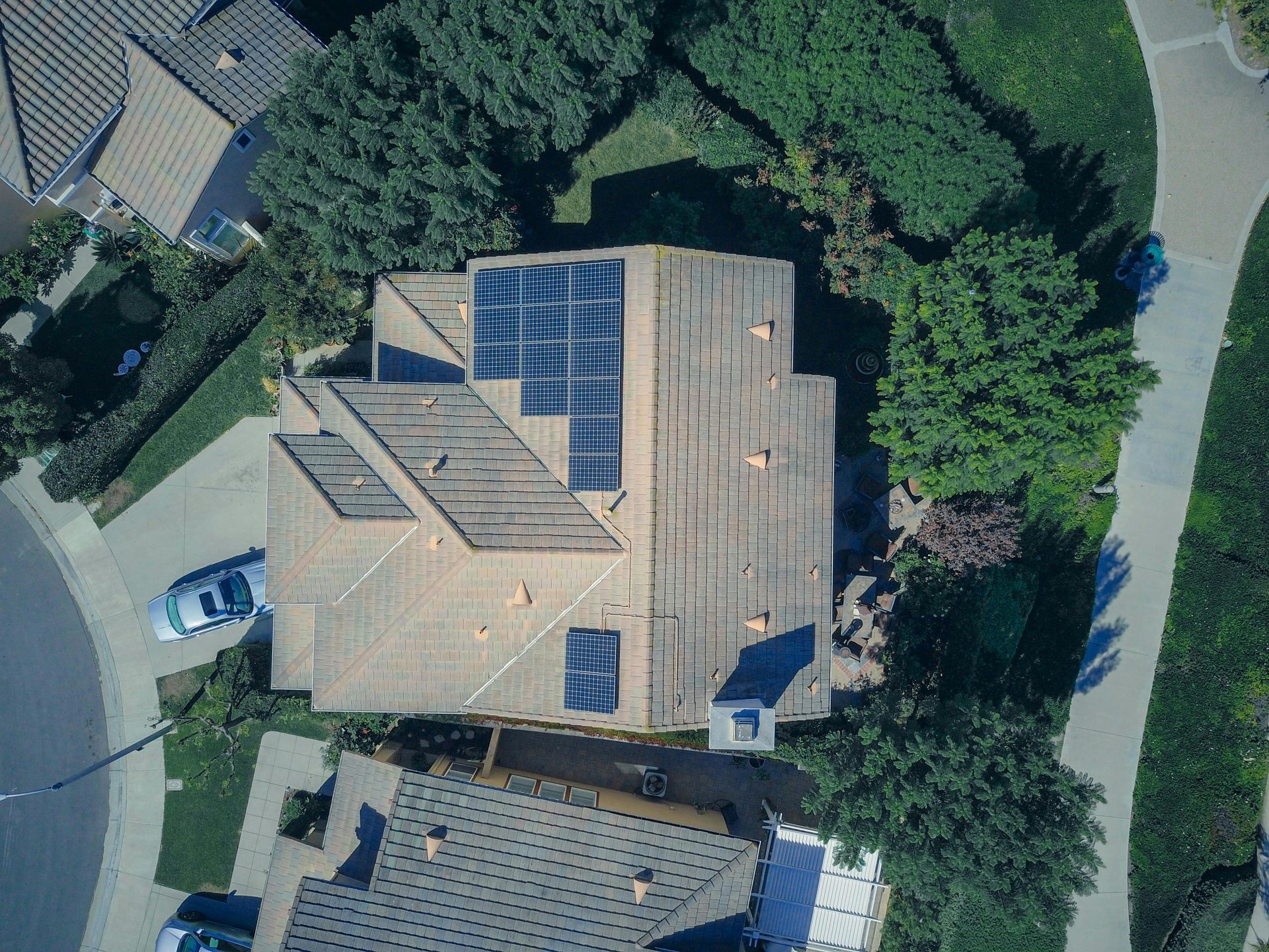 Aerial view of a house with solar panels on the roof, surrounded by trees, a sidewalk, and neighboring houses.