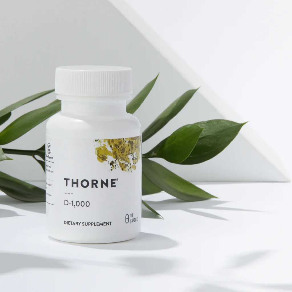 White bottle of Thorne D-1,000 vitamin D supplement, next to green leaves, on a white surface.