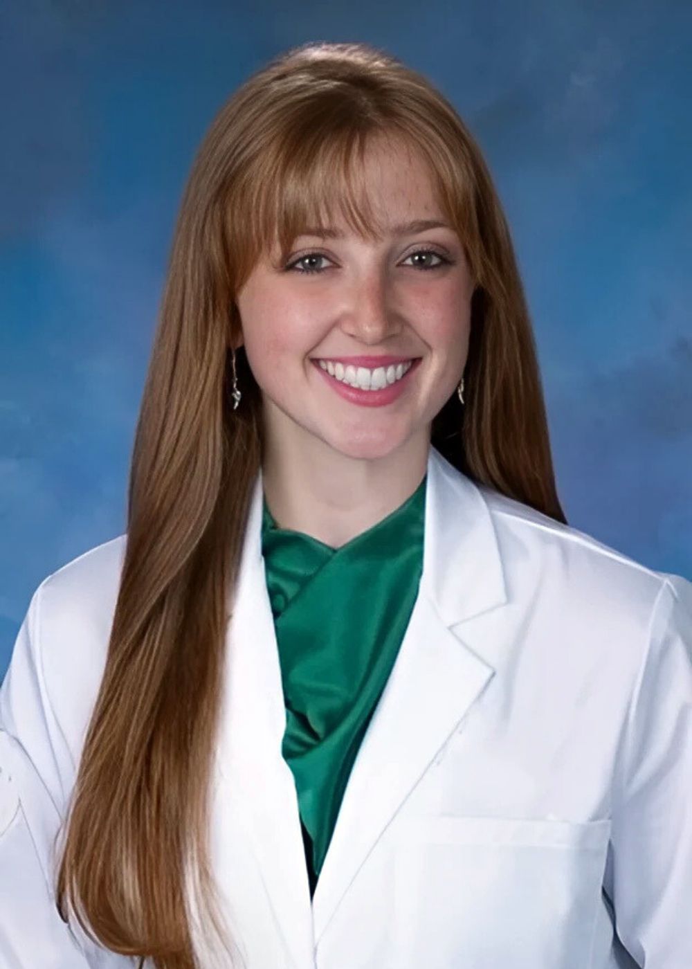 Woman with long red hair, wearing a white lab coat and green scarf, smiling at the camera.