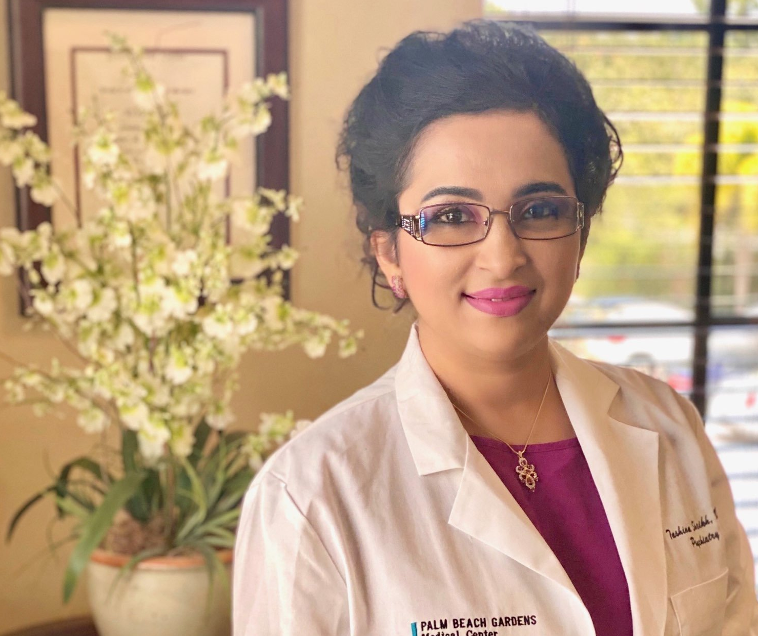 Woman in a white lab coat smiles in a professional setting. Orchid arrangement, framed documents, and window 