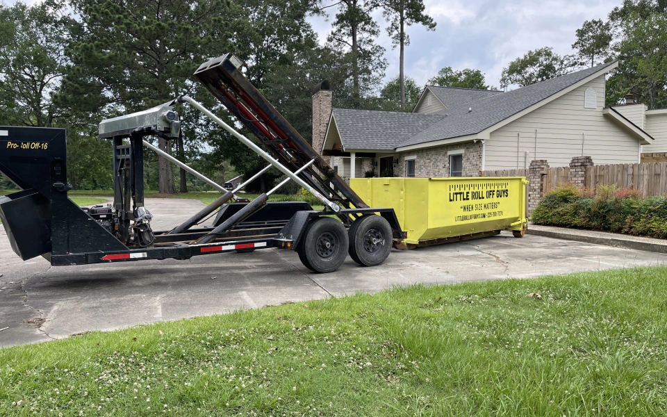 A dumpster is being pulled by a trailer in front of a house.