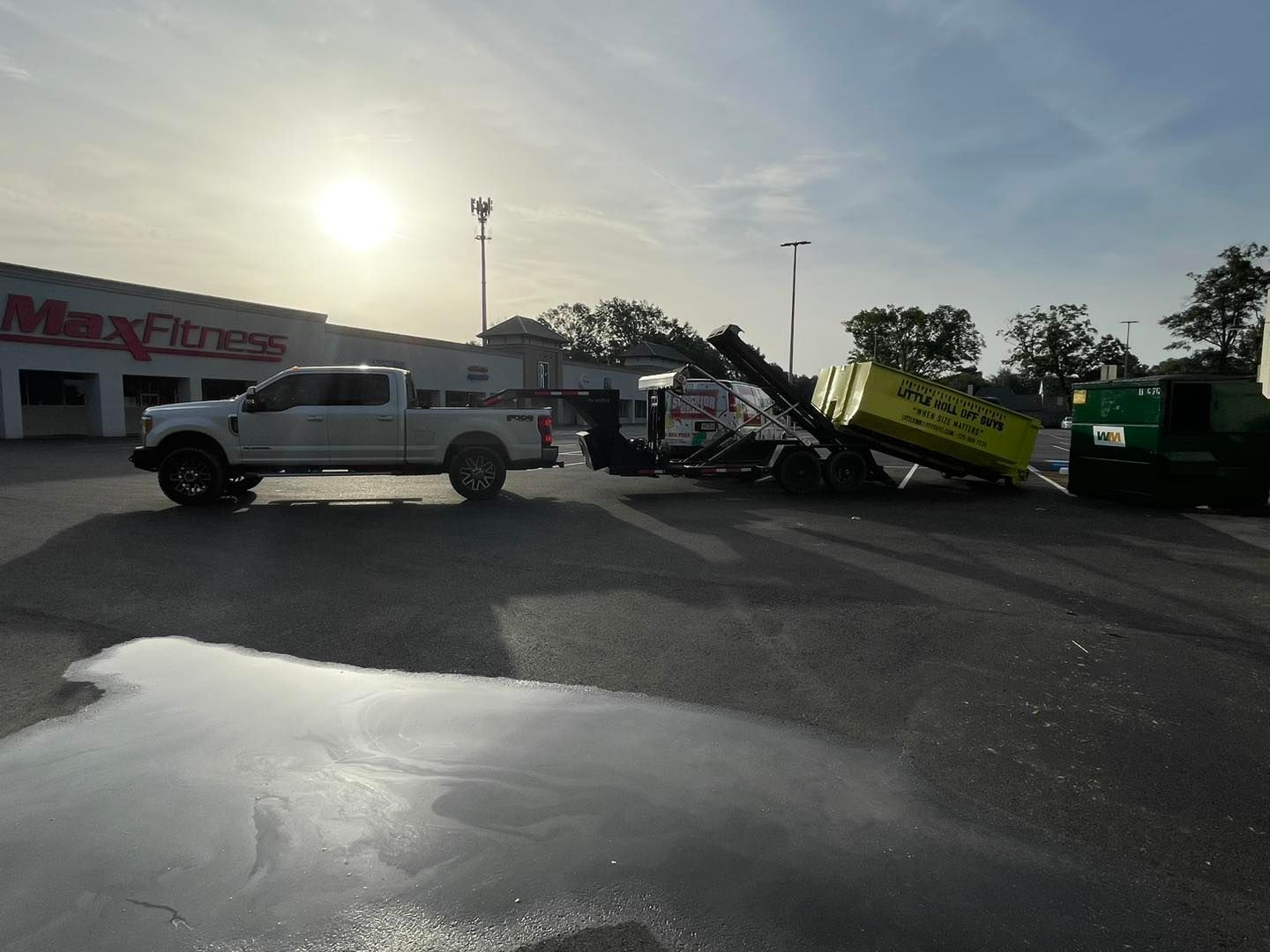 A white truck is towing a yellow dumpster in a parking lot.