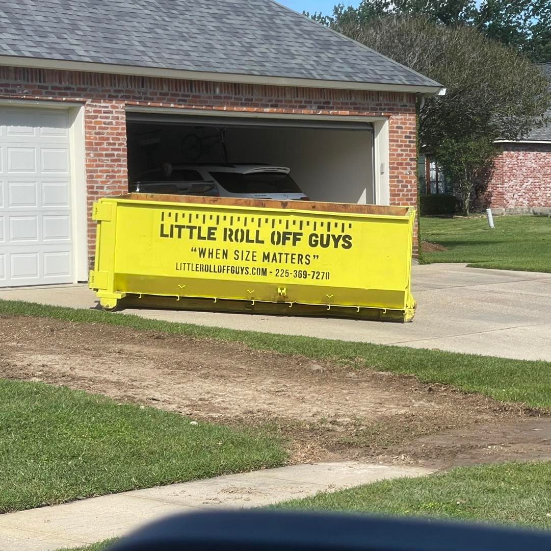 A yellow dumpster that says little roll off guys is in front of a garage.