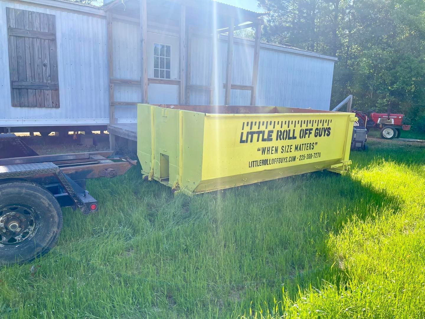 A yellow dumpster is parked in the grass next to a trailer.