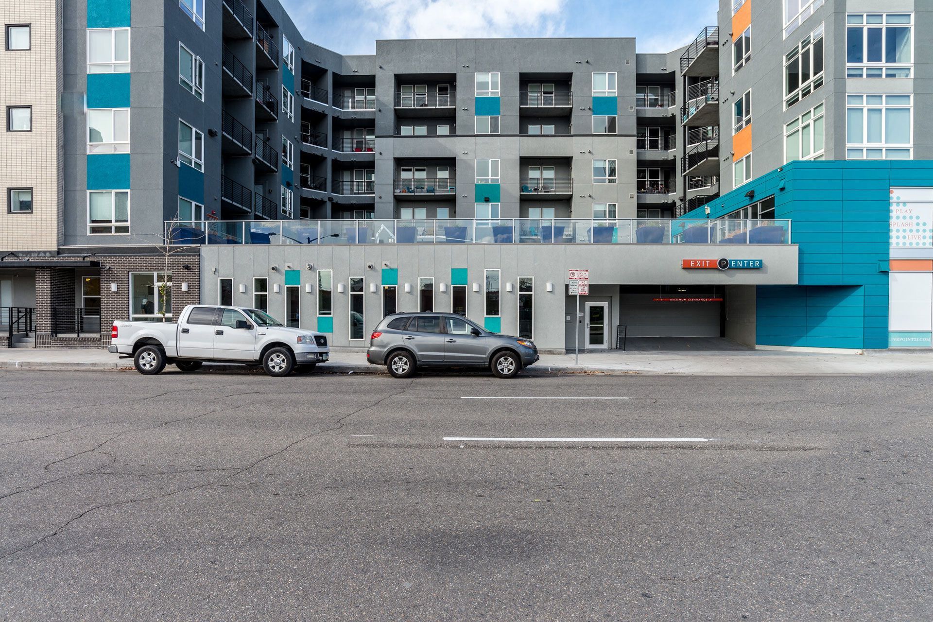 Exterior view of a modern multifamily building with balconies and street-level entrances; two cars parked along the curb.