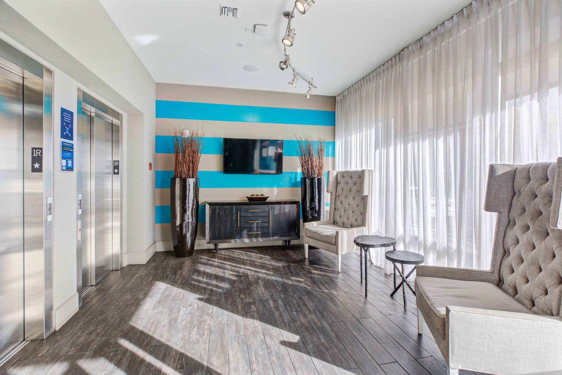 Apartment community lobby with a blue striped wall, two elevators on the left, and lounge seating by large windows.