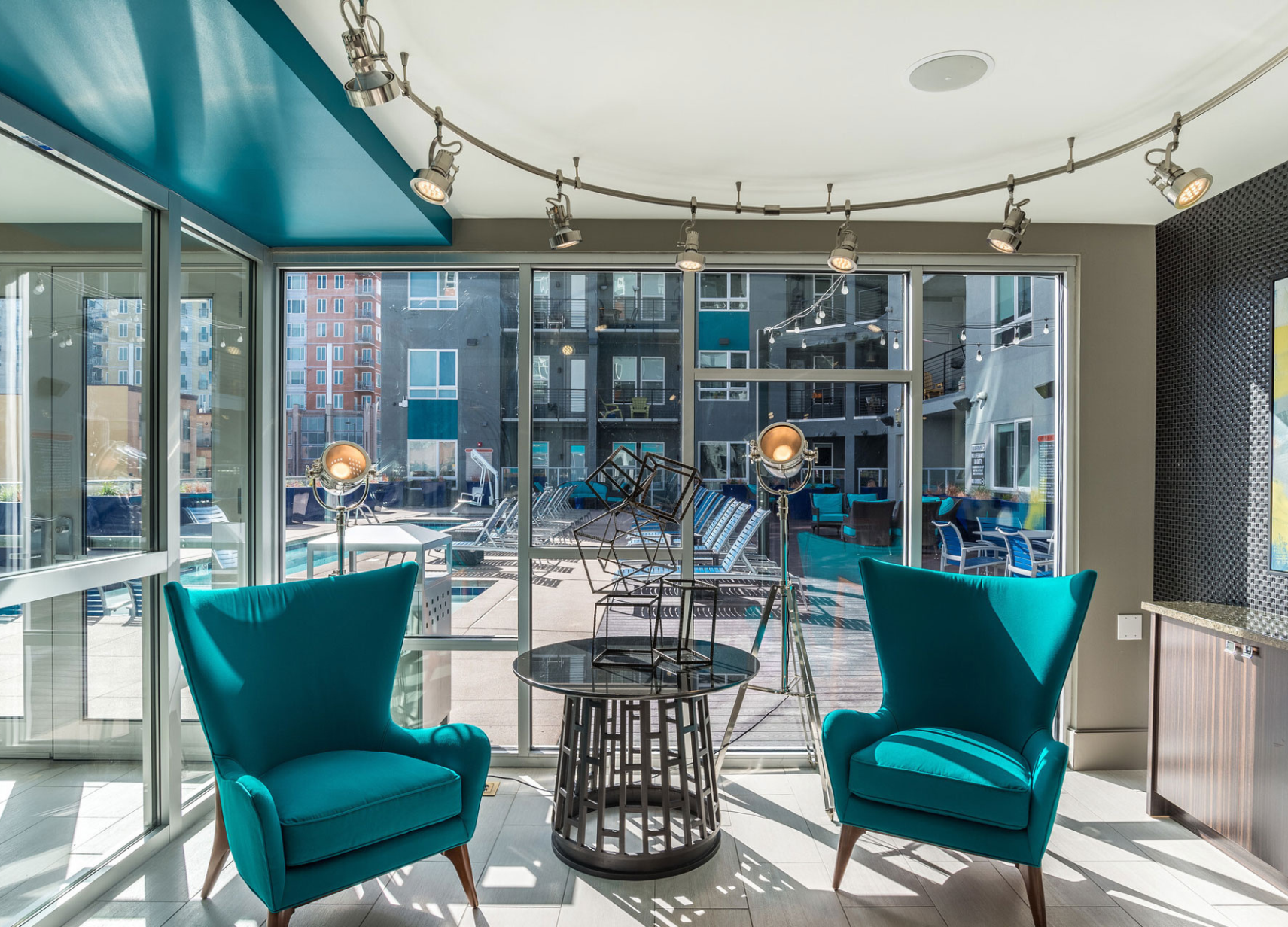 Indoor communal lounge with teal chairs and a round table, view of pool through glass walls.
