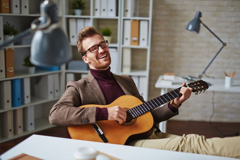 Man in glasses plays guitar, laughing in an office setting, light brown acoustic guitar.
