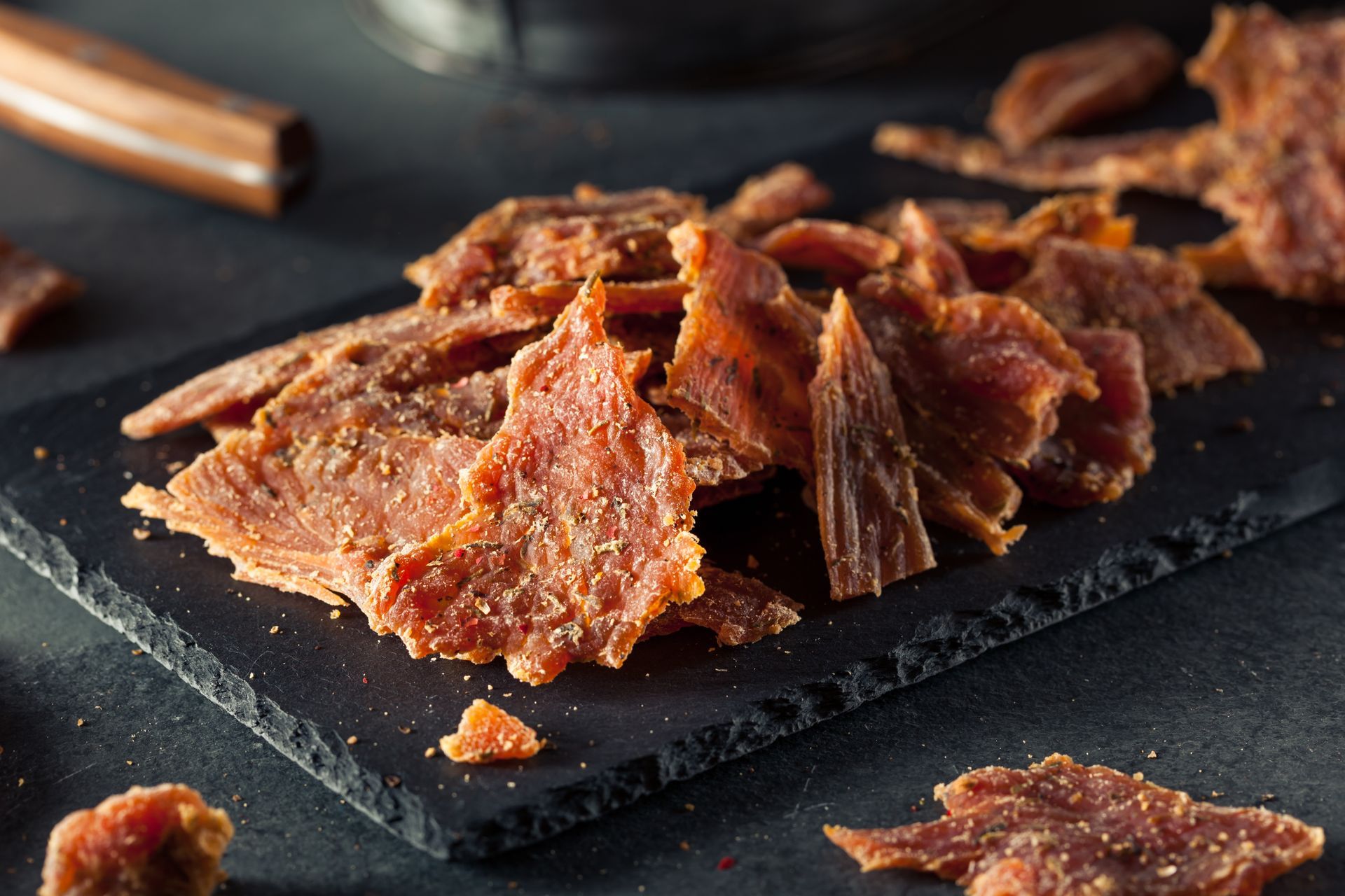 Pile of beef jerky on a dark, textured surface, with a blurred background.