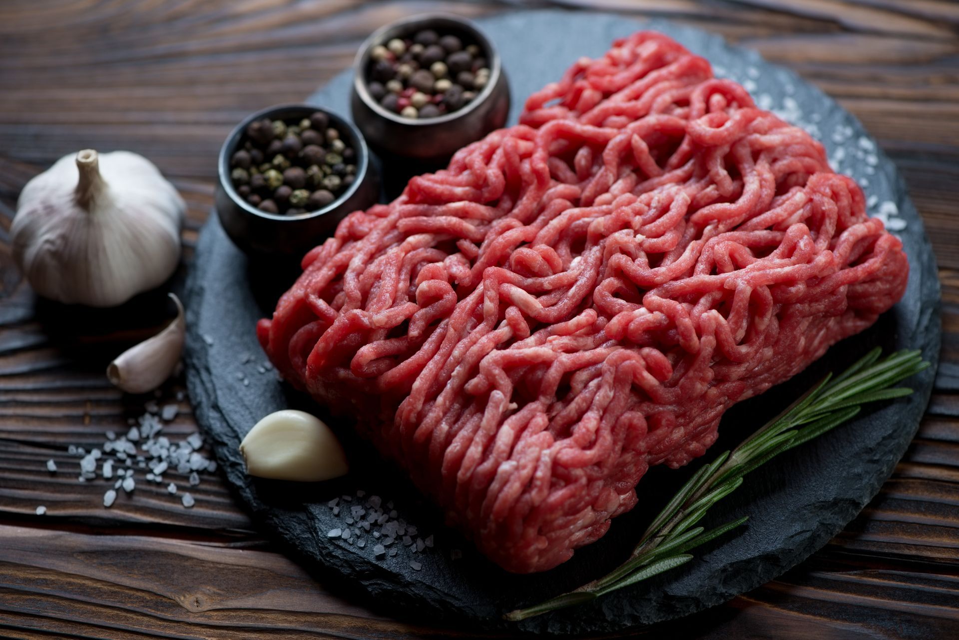Ground beef on a dark slate with peppercorns, garlic, rosemary, and salt on a wooden surface. Ground beef on a dark slate with peppercorns, garlic, rosemary, and salt on a wooden surface.