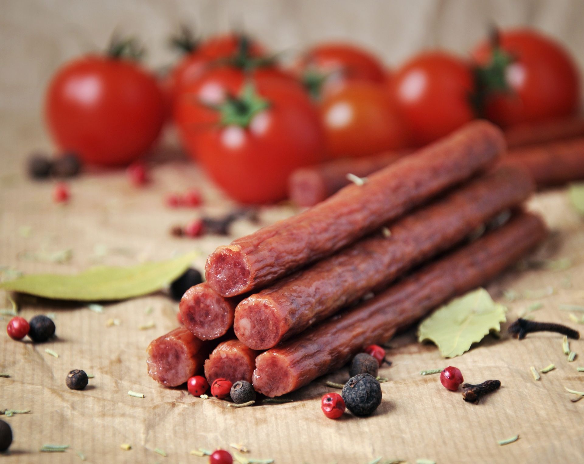Sausage sticks stacked on a paper surface with tomatoes, peppercorns, and herbs in the background.