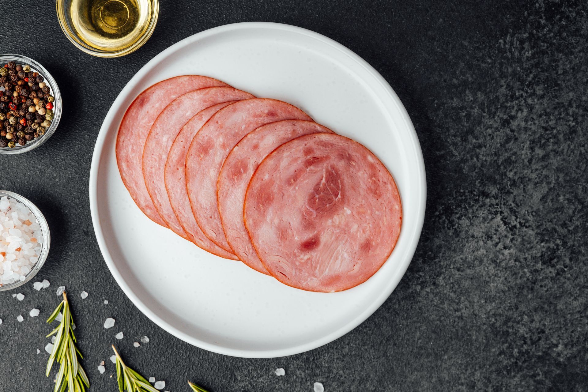 Slices of ham on a white plate, surrounded by spices, oil, and rosemary on a dark surface. Slices of ham on a white plate, surrounded by spices, oil, and rosemary on a dark surface.